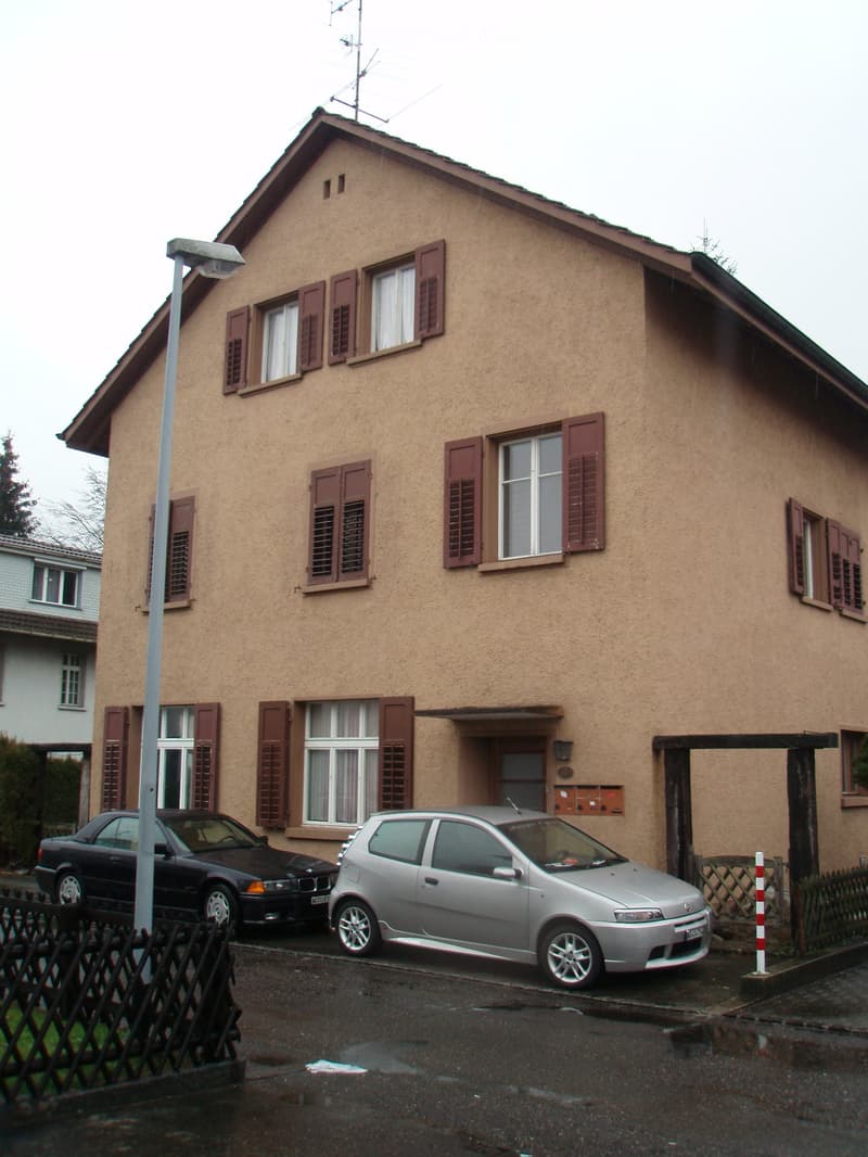 Two-story house with beige siding, multiple windows with brown shutters, and a small entrance area. Two cars are parked in front of the house.