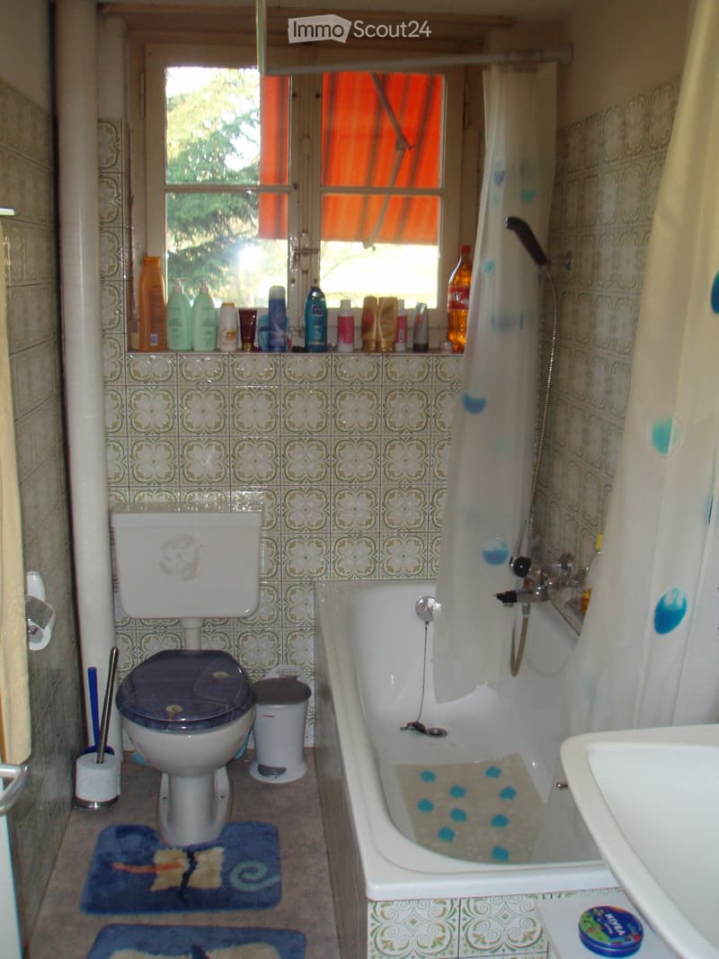Bathroom with a toilet, bathtub, sink, and shower. The bathtub has blue bath salts in it. There is a window with an orange curtain, and various toiletries are placed on the windowsill.