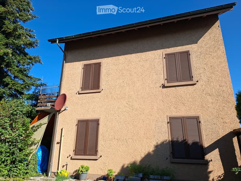 Two-story house with beige stucco exterior, brown shutters, and a small balcony with a satellite dish. Potted plants are placed near the entrance.