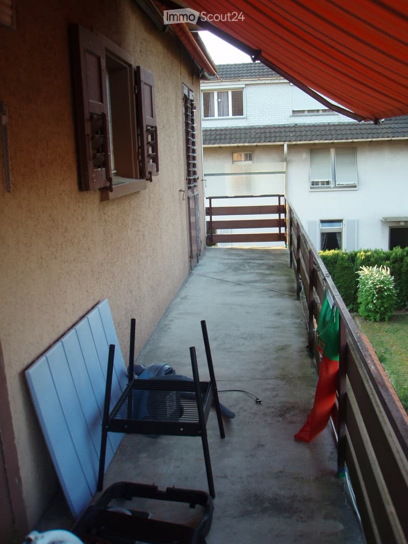 Balcony with a metal railing, a fan, and a screen, with a red awning above and a neighboring building visible in the background.