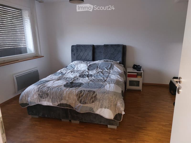 Bedroom with wooden flooring, a large bed with a grey headboard and patterned duvet, a white nightstand with a lamp, and a radiator under the window.