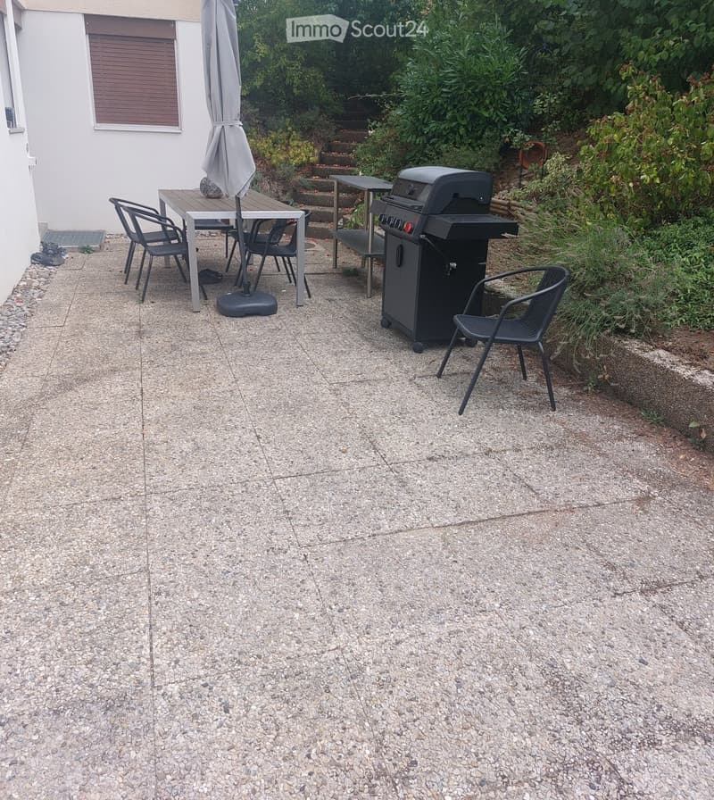 Outdoor patio with a stone floor, a table with chairs, an umbrella, and a grill, surrounded by greenery and a staircase leading up.