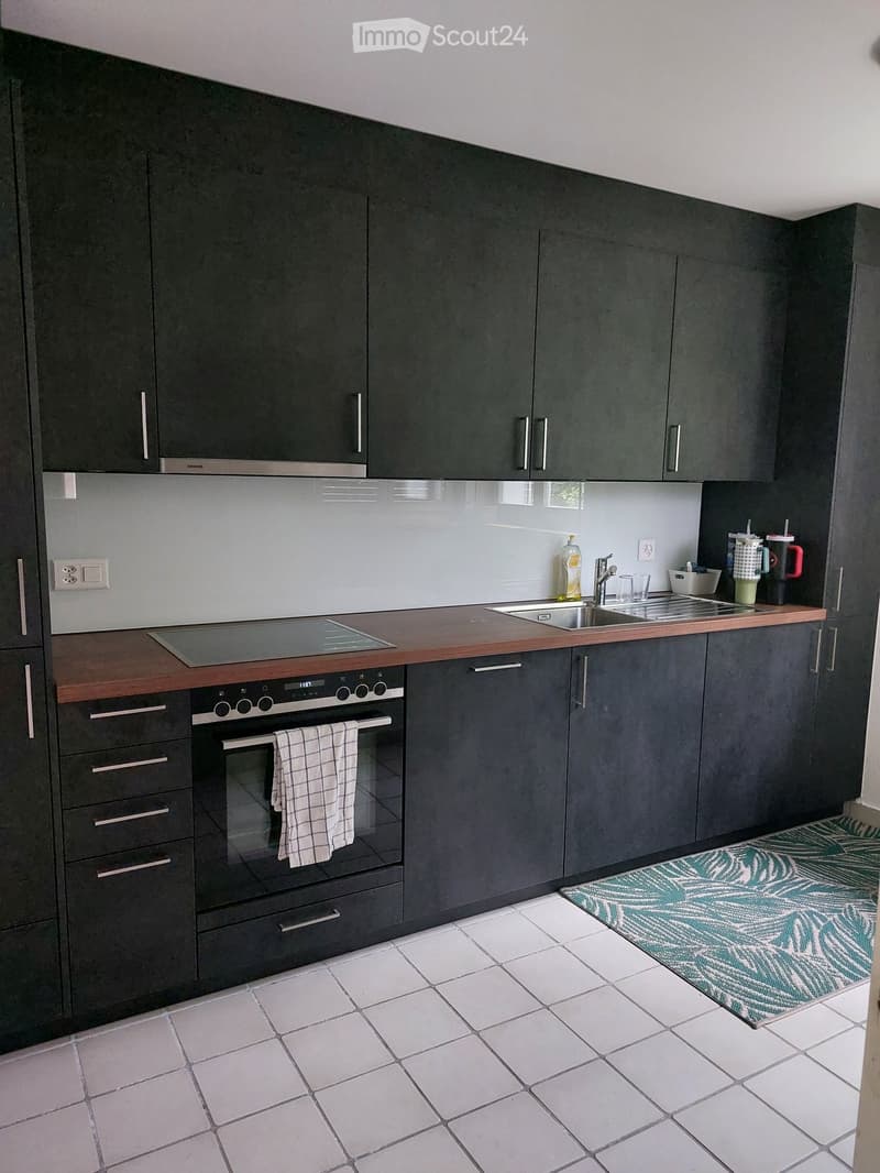 Modern kitchen with dark grey cabinets, white tiled floor, wooden countertop, built-in oven, sink, and various small appliances and items on the counter.