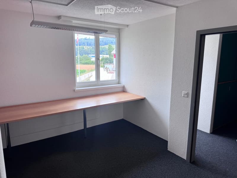 A small, unfurnished room with a single wooden desk, a window with a view of the countryside, and a grey carpeted floor.