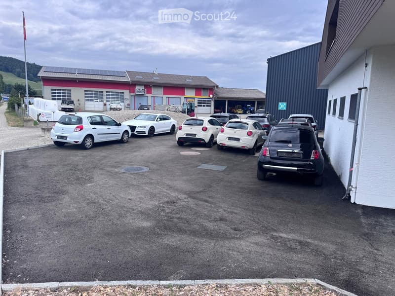 Parking lot with several parked cars, adjacent to a building with garage doors and solar panels on the roof.