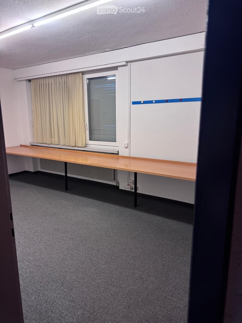 Empty room with grey carpet, wooden desk, window with blinds, and beige curtains.