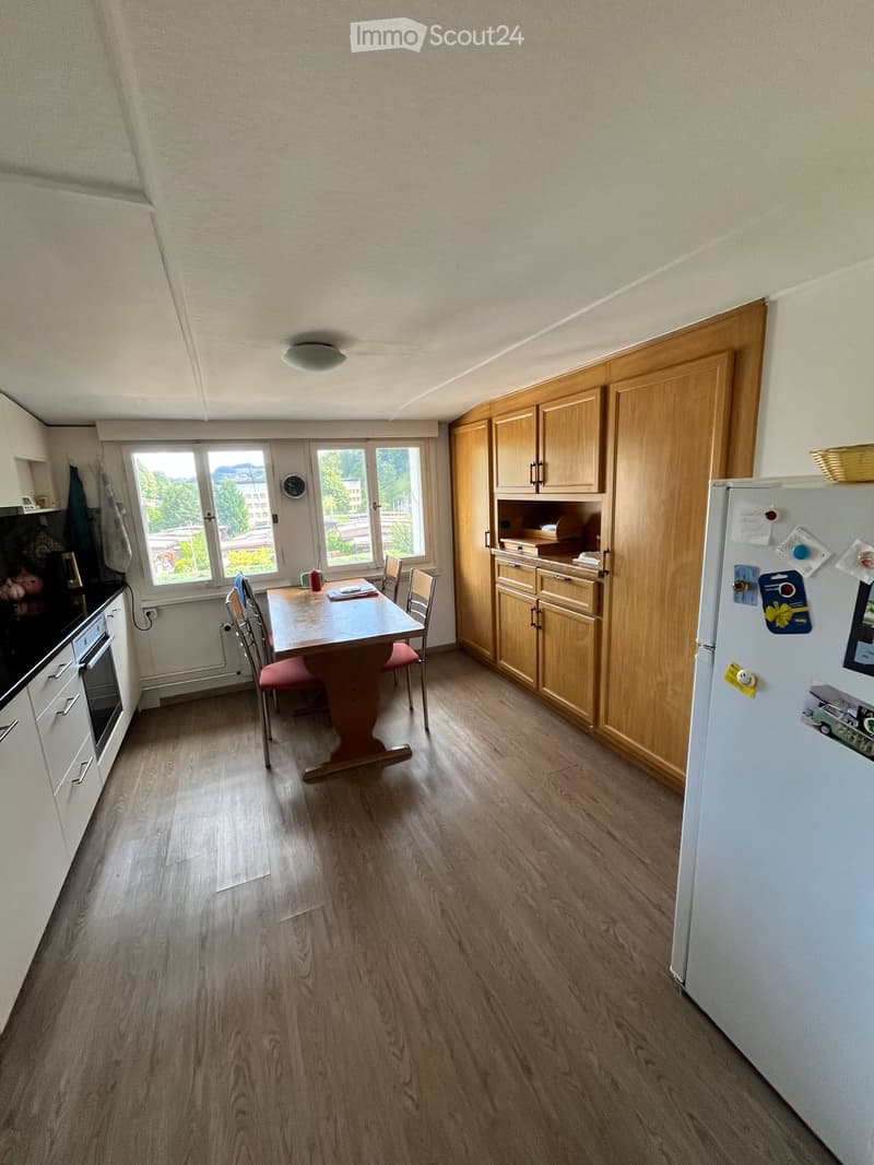 Bright kitchen with wooden cabinets, a dining table with chairs, and a refrigerator. Large windows provide natural light and a view of the outdoors.