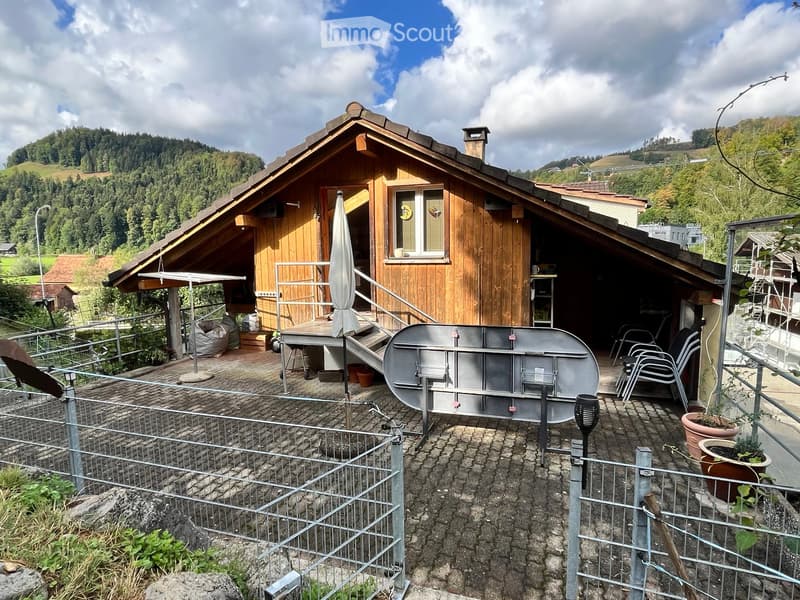 Wooden house with a balcony, outdoor seating area, and a large metal hot tub. The area is surrounded by a metal fence and features potted plants and a scenic mountain view.