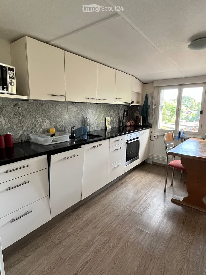 Modern kitchen with white cabinets, black countertops, and a wooden floor. Features a microwave, oven, sink, and a small dining area with a table and chair.