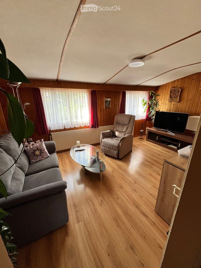 Living room with wooden flooring, large window with curtains, sofa, armchair, coffee table, TV stand, and decorative items.