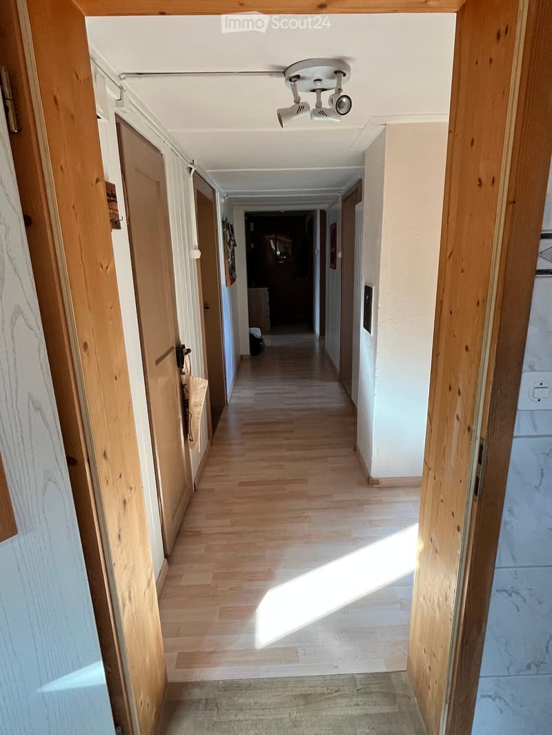 A long, well-lit hallway with wooden flooring and white walls, featuring several doors and a ceiling light fixture.