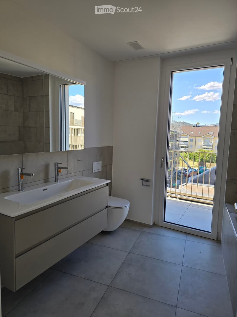 Modern bathroom with a large sink, toilet, and mirror, opening to a balcony with a view of the city.