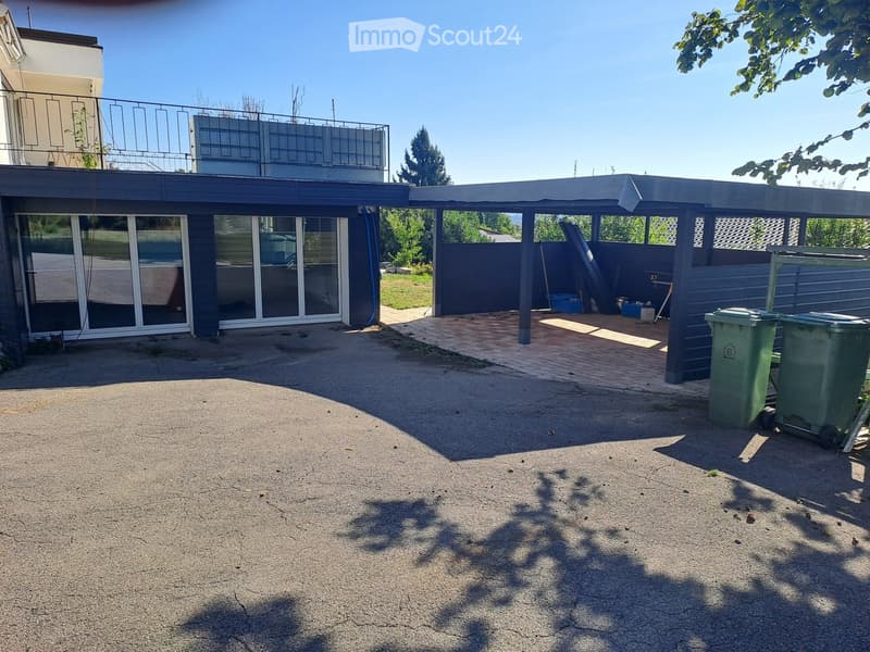 Driveway area in front of a modern house with a covered carport, green trash bins, and a partially visible balcony on the upper level.