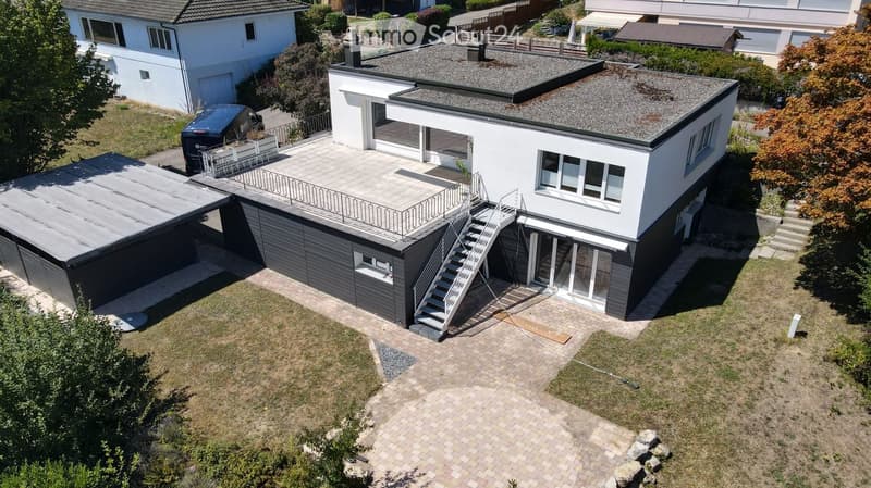 Modern two-story house with a flat roof, garage, and balcony. The house features large windows and a clean, minimalist design.