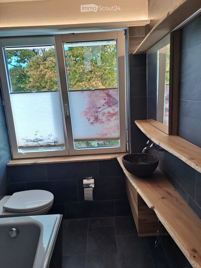 Modern bathroom with dark tiles, a wooden shelf, a black sink, a toilet, and a bathtub. Large windows with blinds offer a view of greenery outside.