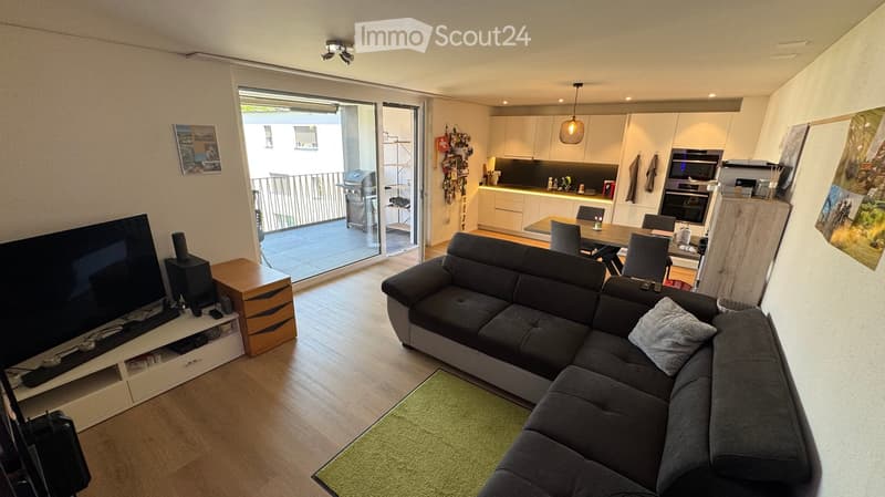 Modern living and dining area with a grey sectional sofa, coffee table, TV stand, dining table with chairs, and a modern kitchen with cabinets, oven, and microwave. The space features wooden flooring, large windows, and a balcony access.