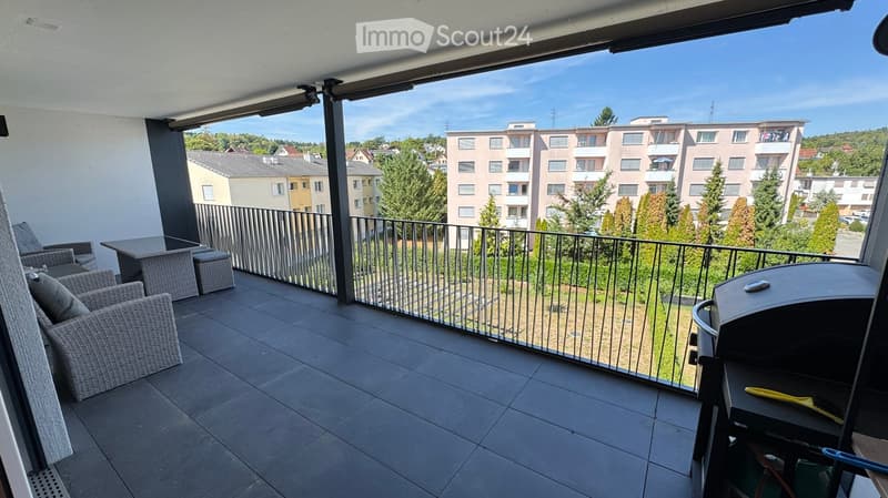 Balcony with tiled floor, metal railing, outdoor furniture including chairs, a table, and a grill, overlooking a residential area with buildings and trees.