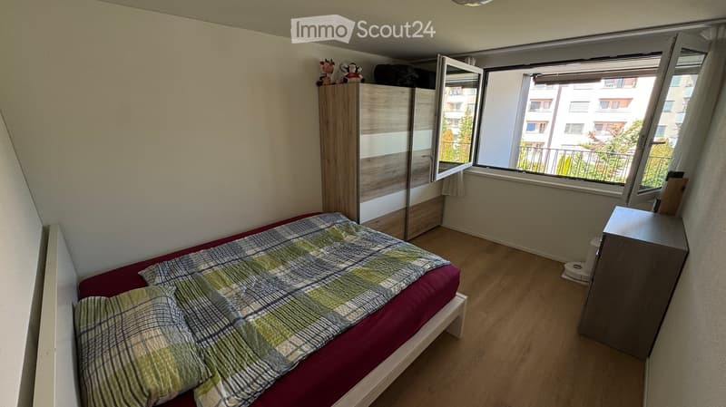 Cozy bedroom with a single bed, wardrobe, nightstand, and mirror. The room features wooden flooring and large windows with a view of the balcony and surrounding buildings.