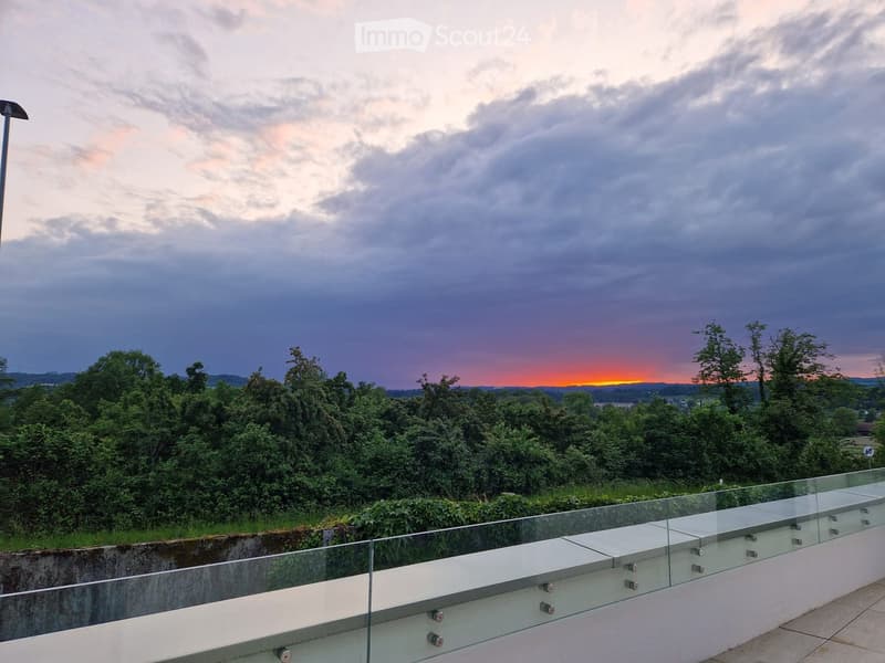 A modern balcony with a glass railing, overlooking a lush green landscape and a dramatic sunset sky.