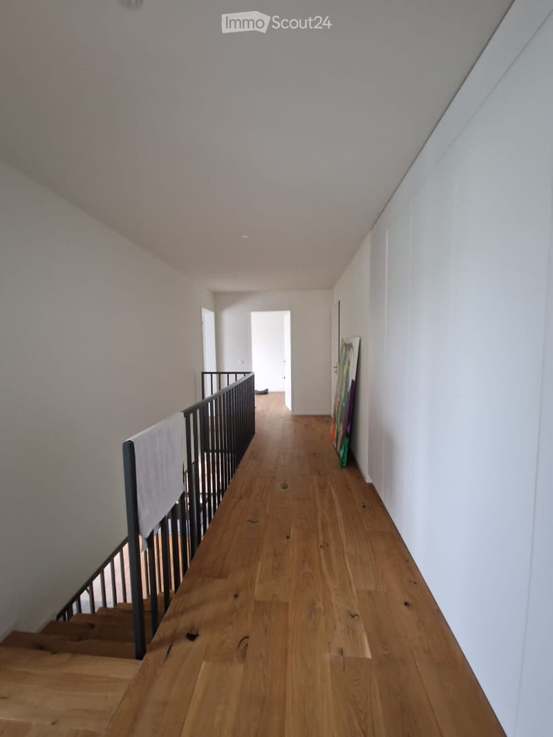 A narrow hallway with wooden flooring, white walls, and a staircase leading upwards. A painting leans against the wall on the right side.