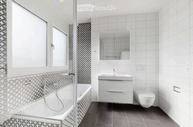 Modern bathroom with white tiles, a bathtub, a sink with a cabinet, a toilet, and a large mirror above the sink. The walls feature a geometric tile pattern in black and white.