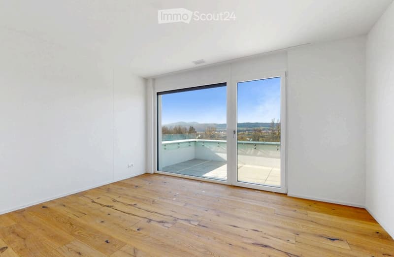 Bright, empty room with wooden flooring and large sliding glass doors opening to a balcony with scenic views.
