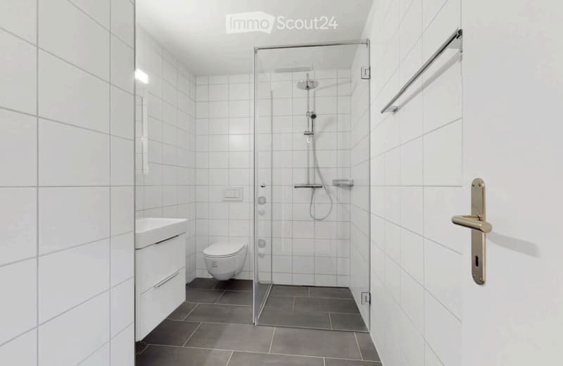 Modern bathroom with white tiled walls and floor, featuring a toilet, sink, glass-enclosed shower, and towel rack.