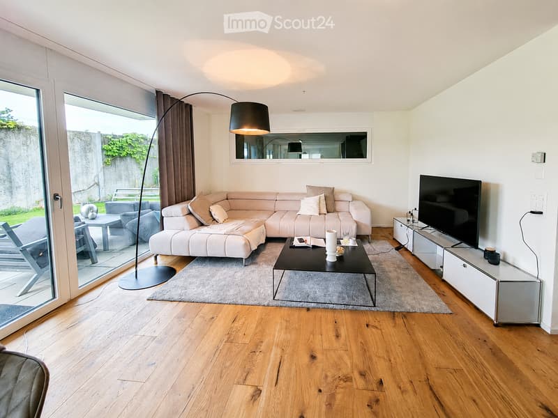 Modern living room with light-colored sofa, coffee table, TV stand, floor lamp, and large windows offering a view of the garden.