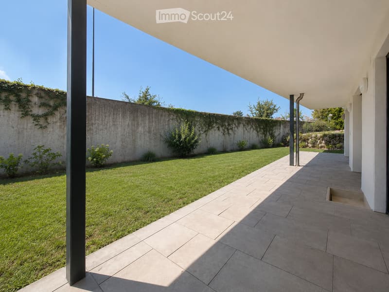 Modern outdoor patio with tiled flooring, covered roof, and a well-maintained lawn surrounded by a low concrete wall and greenery.