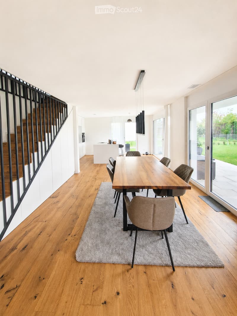Modern dining area with a wooden dining table, six chairs, a gray rug, pendant lights, and a kitchen island visible in the background.