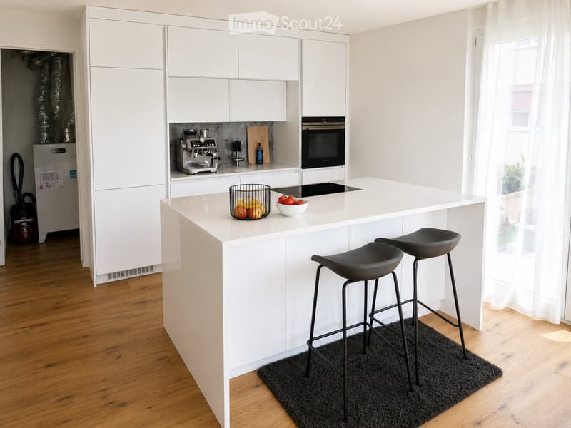 Modern white kitchen with a central island, two black bar stools, coffee maker, oven, refrigerator, and decorative fruit bowls.