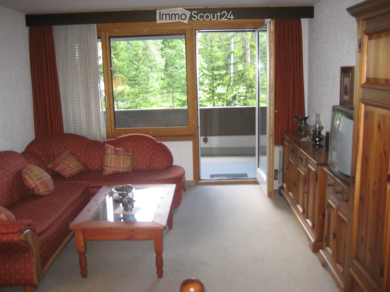 Living room with a red patterned sofa, wooden coffee table, television on a wooden cabinet, and large windows offering a view of greenery.