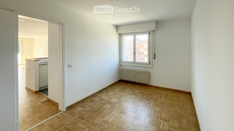 Empty room with light-colored walls, wooden herringbone flooring, and a window with blinds. A radiator is visible below the window, and an open doorway leads to another room.