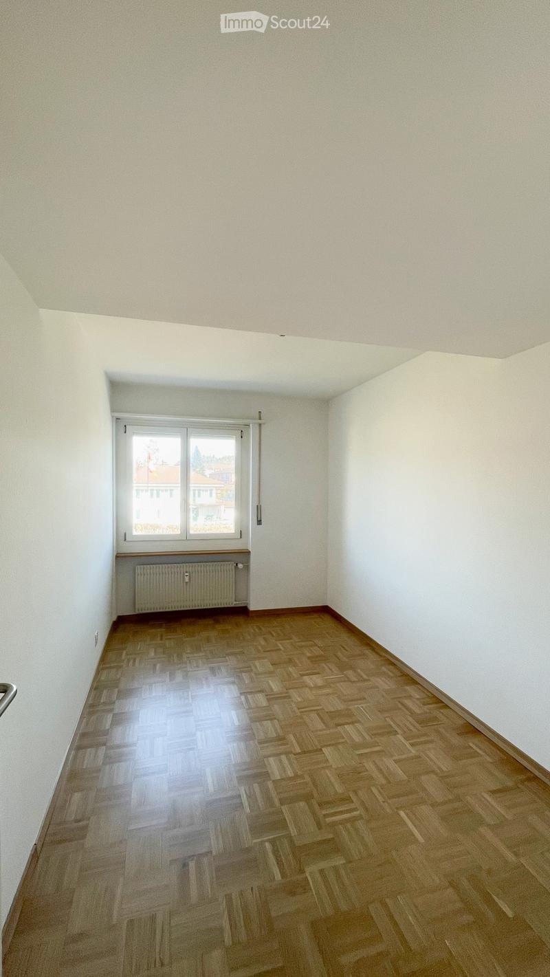 Empty room with wooden herringbone flooring, white walls, and a window with a radiator below it.