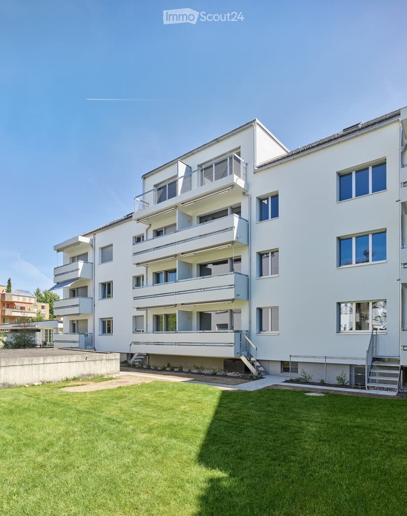 Modern multi-story apartment building with white facade, multiple balconies, and a well-maintained lawn in front.