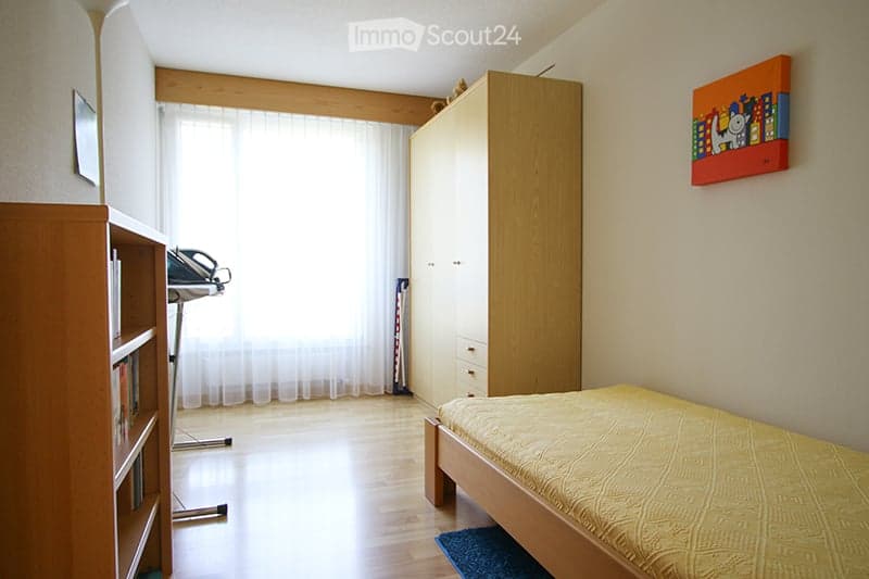 Bright bedroom with wooden flooring, a single bed with yellow bedding, a large wardrobe, a bookshelf, and an exercise bike near the window.