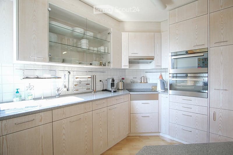 Modern kitchen with light wood cabinets, stainless steel appliances, and a clean, organized design. Features include a sink, faucet, oven, dishwasher, coffee maker, teapot, and dish rack.