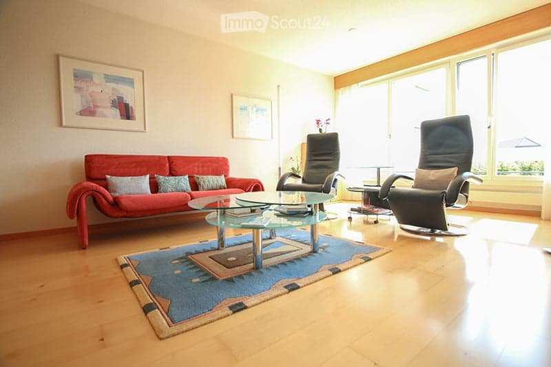 Bright living room with a red sofa, glass coffee table, two black chairs, and a patterned rug on a wooden floor.