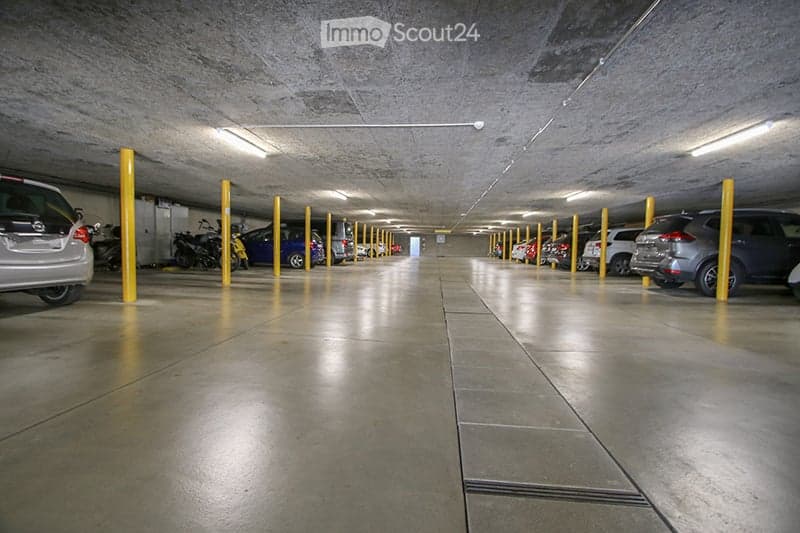Underground parking area with multiple parked cars and motorcycles, concrete floors and walls, yellow support pillars, and fluorescent lighting.