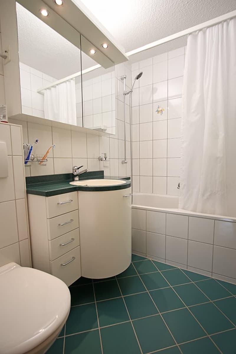 Modern bathroom with white tiled walls, green countertop, round sink cabinet, bathtub, showerhead, and a large mirror above the sink.