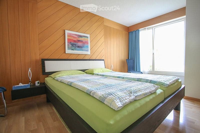 Bright bedroom with wooden walls and flooring, featuring a double bed with green bedding, a nightstand with a lamp, a chair, and a picture frame on the wall.