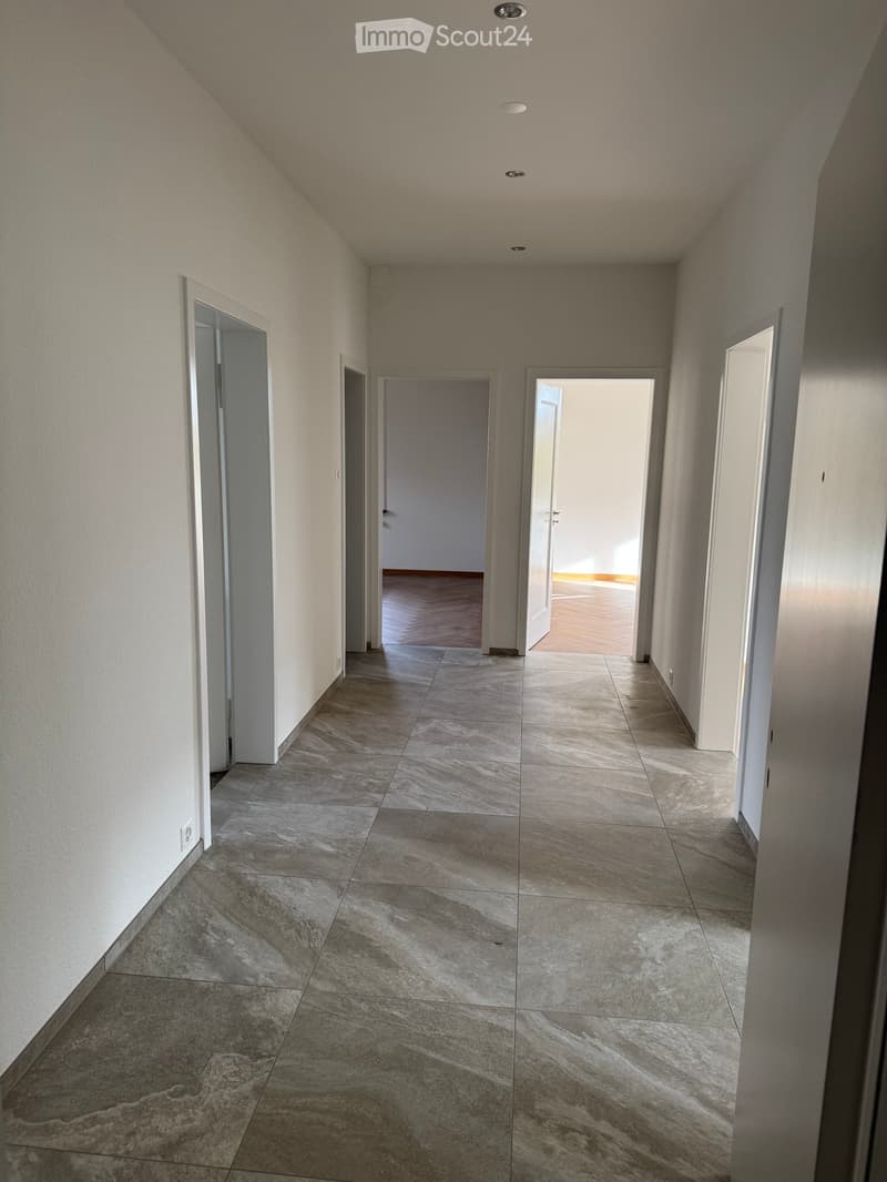 Empty corridor with white walls, wooden floor, tiled floors, two closed doors, one open door leading to a room with light.