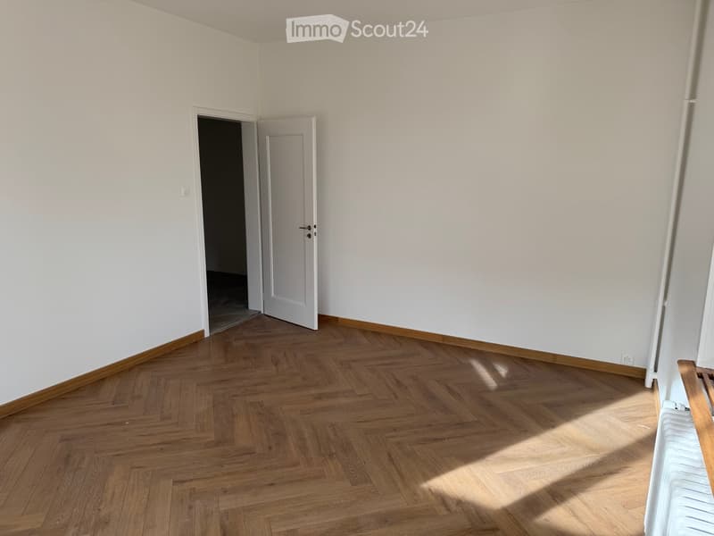 Empty room, wooden parquet flooring, white walls, 2 doors, one leading to another room, wooden bench against the wall