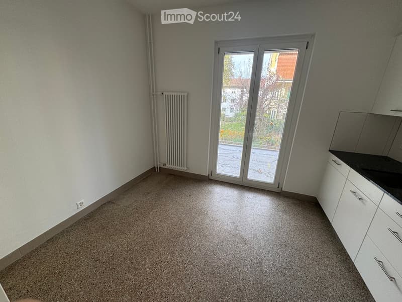 Empty room, grey floor, white walls, large sliding glass door, radiator, electrical outlet, kitchen area with white cabinets and black countertops