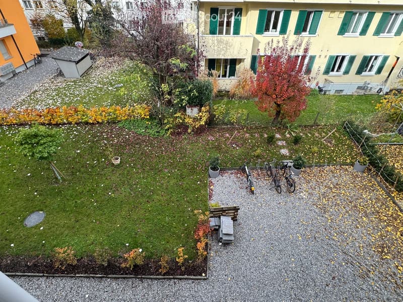Green garden with gravel path, surrounded by bushes, trees, and fallen leaves. Bicycles are parked on gravel near potted plants.