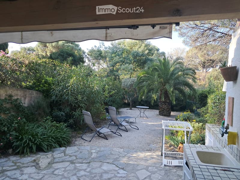 Outdoor garden area with stone-paved pathway, lounge chairs, small tables, lush greenery, and a sink area.