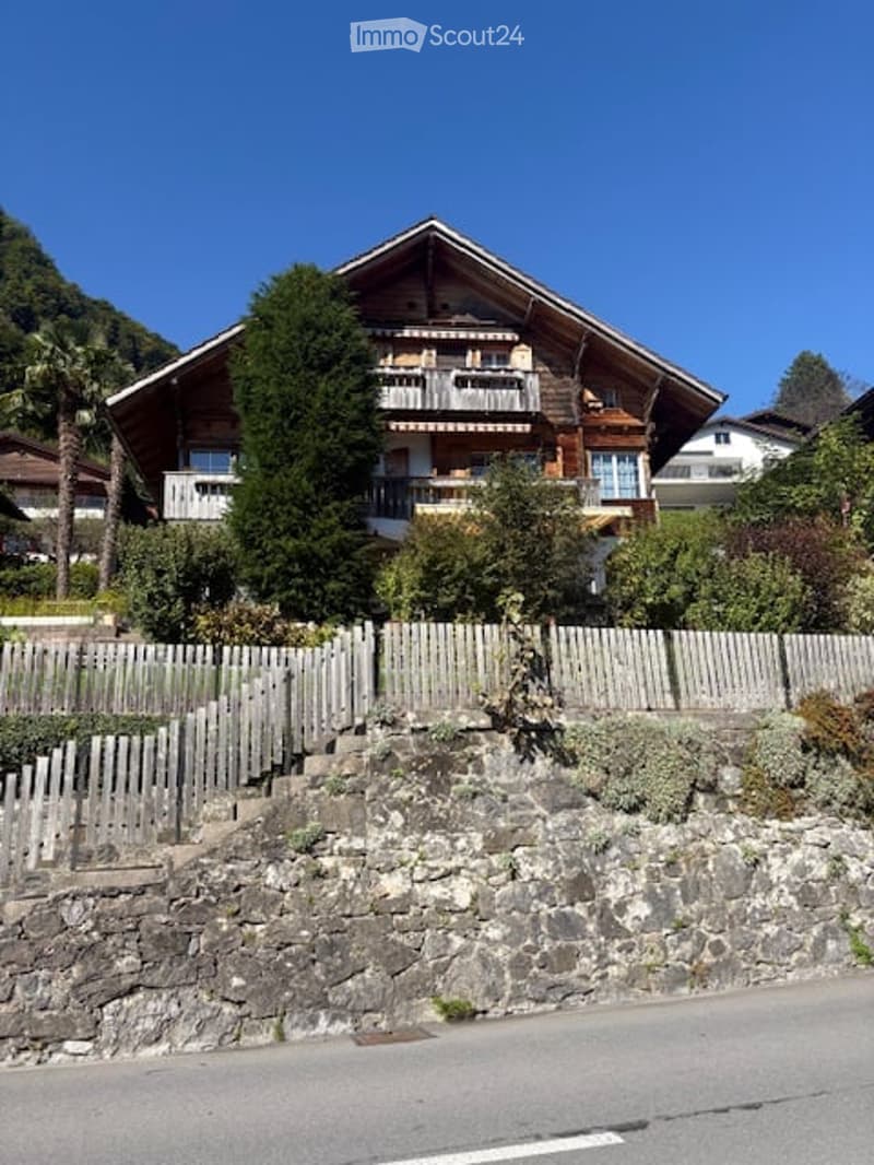 A two-story wooden house with balconies, surrounded by greenery and a stone wall.