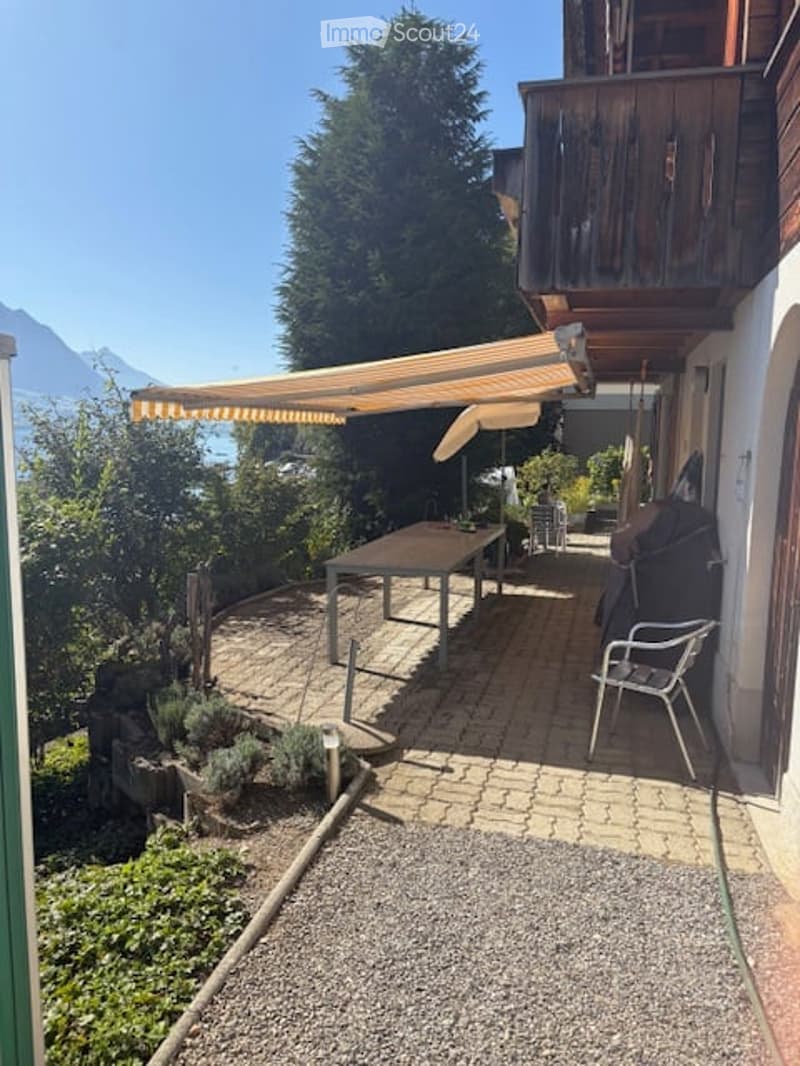 Outdoor patio with a wooden table, chairs, and a retractable awning, surrounded by greenery and offering a scenic mountain view.