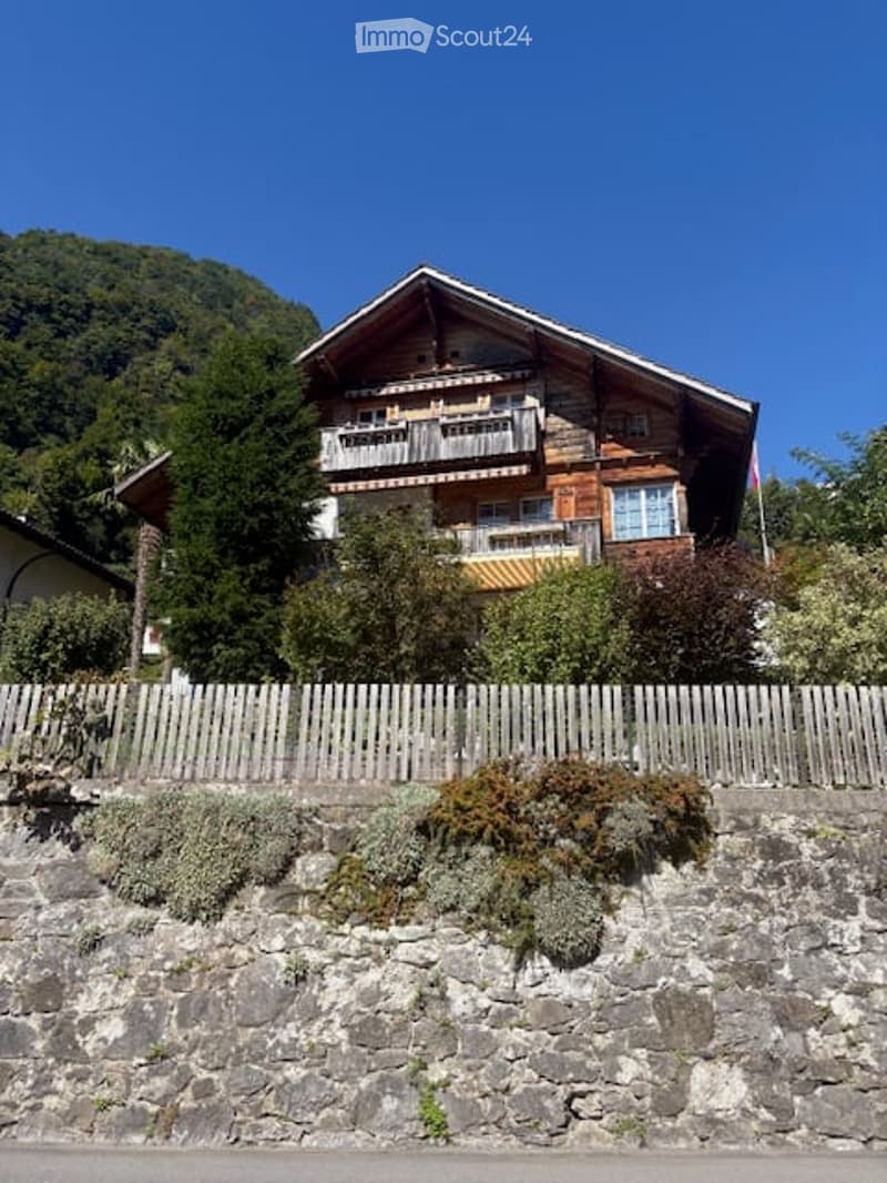 A two-story wooden house with balconies, surrounded by greenery and a stone wall.