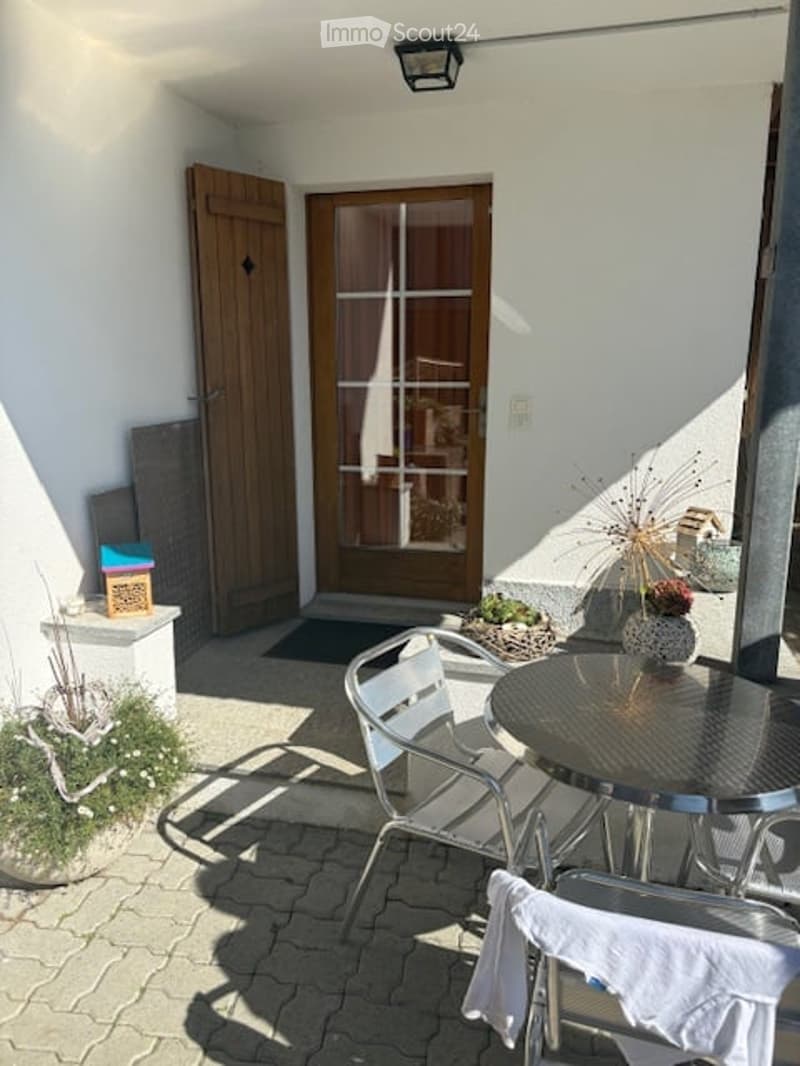 Balcony with a round metal table, two white chairs, a vase with dried plants, and a small potted plant. Wooden door and white walls are visible in the background.